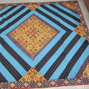 May include: A large, colourful textile with a geometric pattern. The design features a central diamond of orange and yellow squares, surrounded by blue and black striped sections. The border is a repeating pattern of red, yellow, and purple shapes.