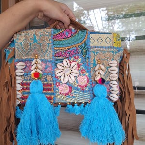 Può includere: Borsa a mano patchwork colorata con base turchese, ornata con conchiglie, nappe blu e frange marroni. La borsa presenta ricami floreali e dettagli dorati, creando un'estetica bohémien. Il testo "vintagebynihal" è visibile.