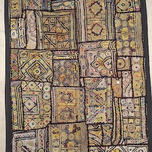 Op de afbeelding: Een rechthoekig wandkleed bestaande uit een patchwork van geborduurde stoffen panelen in goud-, geel- en bruintinten. Het textiel is versierd met ingewikkelde geometrische patronen, metaaldraadstiksels en kleine ronde spiegeltjes.