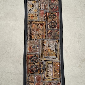 May include: A long, rectangular tapestry with a black border and a patchwork design. The tapestry is made of various fabrics in shades of brown, gold, and red, and is decorated with sequins and small mirrors. The tapestry has a vintage feel and is perfect for adding a touch of bohemian style to any room.
