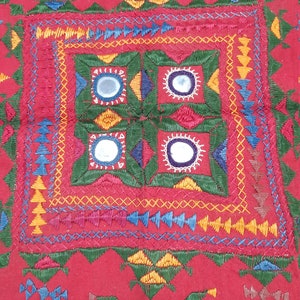 Pu&ograve; includere: Un tessuto rosso vibrante con ricami intricati. Il design presenta motivi geometrici, tra cui quadrati, triangoli e cerchi, in una variet&agrave; di colori come verde, giallo, blu e bianco. Piccoli specchi sono incorporati nel design.