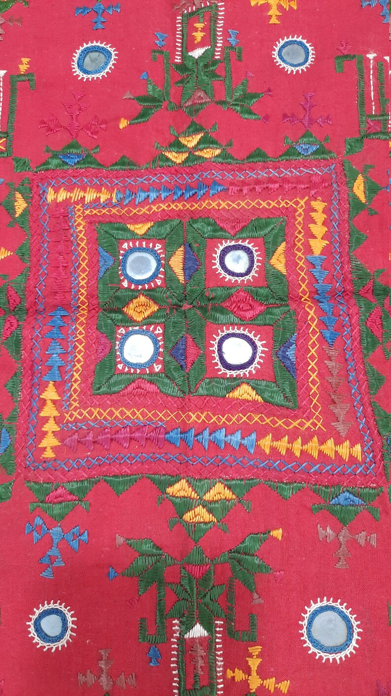 Pu&ograve; includere: Un tessuto rosso vibrante con ricami intricati. Il design presenta motivi geometrici, tra cui quadrati, triangoli e cerchi, in una variet&agrave; di colori come verde, giallo, blu e bianco. Piccoli specchi sono incorporati nel design.