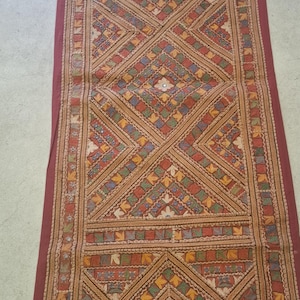 Puede incluir: Textil rectangular largo con un patrón geométrico en tonos rojos, beige, verdes y azules. El diseño central presenta formas de diamante y motivos florales, con un borde rojo oscuro. El textil tiene una estética vintage.