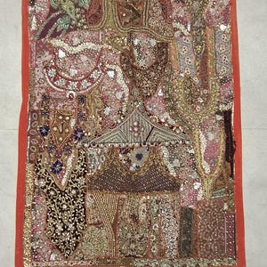 May include: A decorative textile, rectangular in shape, with a red border. The central design is a patchwork of fabrics in red, brown, gold, and green, adorned with beads and sequins. The overall appearance is rich and detailed.