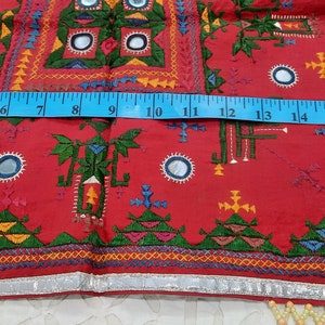 Pu&ograve; includere: Un tessuto rosso vibrante con ricami intricati e abbellimenti a specchio. Il design presenta motivi geometrici in verde, giallo, blu e arancione. Un metro a nastro blu mostra una lunghezza di circa 45 cm. I bordi sono rifiniti con tessuto argentato e frange di perline.