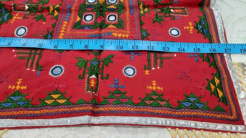 Pu&ograve; includere: Un tessuto rosso vibrante con ricami intricati e abbellimenti a specchio. Il design presenta motivi geometrici in verde, giallo, blu e arancione. Un metro a nastro blu mostra una lunghezza di circa 45 cm. I bordi sono rifiniti con tessuto argentato e frange di perline.