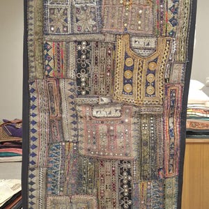 Puede incluir: Textil de patchwork con borde negro, con cuadrados de tela bordados y espejados. El diseño incorpora azules, rosas y dorados, creando una pieza decorativa texturizada.
