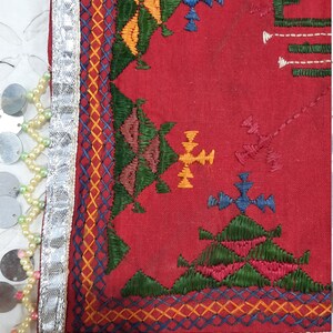 Pu&ograve; includere: Primo piano di un tessuto rosso con ricami intricati in verde, giallo, blu e arancione. Il bordo &egrave; rifinito con tessuto argentato, piccoli dischi metallici e fili di perline. Un piccolo specchio &egrave; incastonato nel tessuto.