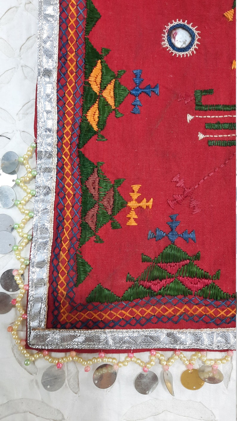Pu&ograve; includere: Primo piano di un tessuto rosso con ricami intricati in verde, giallo, blu e arancione. Il bordo &egrave; rifinito con tessuto argentato, piccoli dischi metallici e fili di perline. Un piccolo specchio &egrave; incastonato nel tessuto.