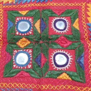 Pu&ograve; includere: Primo piano di un tessuto rosso con ricami intricati. Il design presenta un quadrato centrale con dettagli a specchio, circondato da cuciture verdi, gialle, blu e bianche. I motivi dettagliati e i colori vivaci creano un'estetica artigianale ricca.