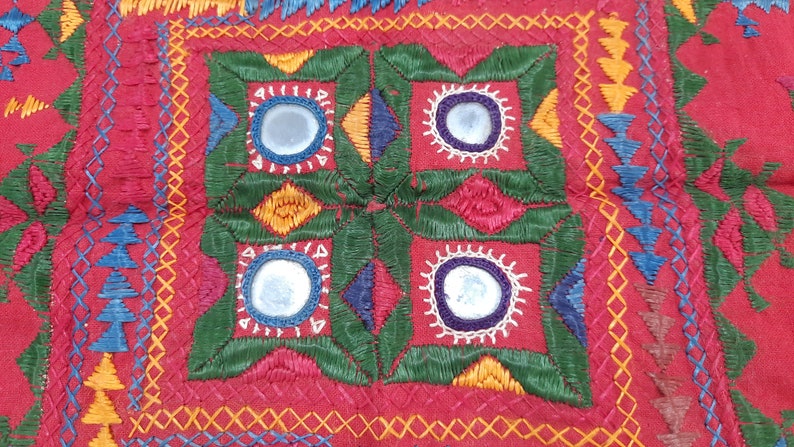 Pu&ograve; includere: Primo piano di un tessuto rosso con ricami intricati. Il design presenta un quadrato centrale con dettagli a specchio, circondato da cuciture verdi, gialle, blu e bianche. I motivi dettagliati e i colori vivaci creano un'estetica artigianale ricca.