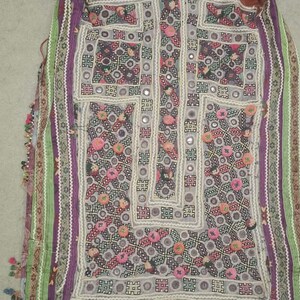 May include: A vintage, intricately embroidered textile in shades of purple, green, and red. The garment features detailed geometric patterns, small mirrors, and colorful floral accents. The edges are trimmed with tassels.