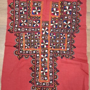 May include: A rectangular red fabric panel featuring an intricate central embroidery design. The pattern consists of geometric shapes, circular motifs, and small reflective mirrors arranged in a symmetrical, multi-colored layout.