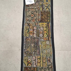 May include: A rectangular, patchwork textile with a black border. The design features various geometric patterns in shades of gold, brown, and cream, with small mirrored accents. The textile has a vintage aesthetic.