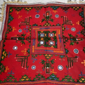 Pu&ograve; includere: Un vivace tessuto rosso con ricami intricati e dettagli a specchio. Il design presenta motivi geometrici in verde, giallo, blu e bianco. I bordi sono rifiniti con tessuto argentato e piccoli abbellimenti rotondi. Si tratta di un tessuto culturale.
