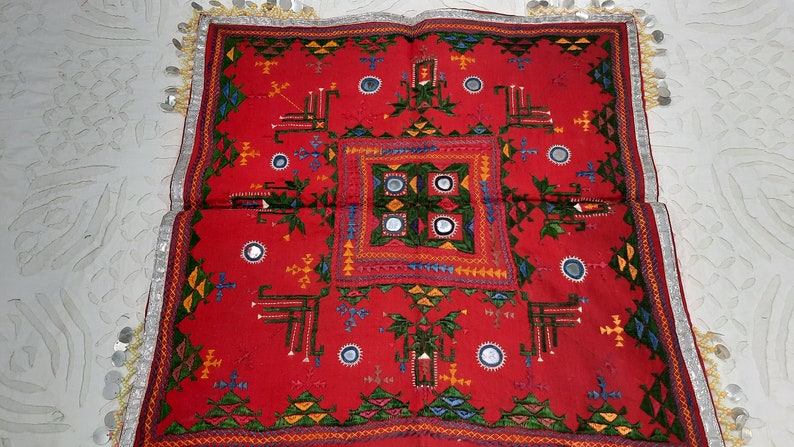 Pu&ograve; includere: Un vivace tessuto rosso con ricami intricati e dettagli a specchio. Il design presenta motivi geometrici in verde, giallo, blu e bianco. I bordi sono rifiniti con tessuto argentato e piccoli abbellimenti rotondi. Si tratta di un tessuto culturale.
