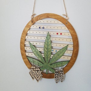 Könnte beinhalten: Ein Ohrringhalter aus Holz in Form eines Marihuana-Blattes mit einem braunen Holzrahmen. Der Halter hat mehrere Löcher für Ohrringe und ist mit bunten Strasssteinen verziert. Zwei Paar Ohrringe hängen am Halter.