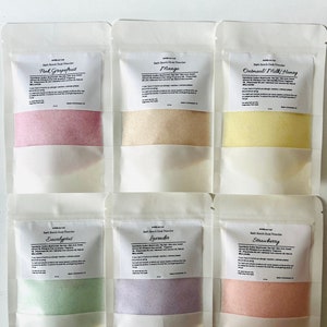 May include: Six small white pouches with clear plastic windows, each containing a different colored bath bomb dust powder. The pouches are labeled with the names of the scents: Pink Grapefruit, Mango, Oatmeal Milk Honey, Eucalyptus, Lavender, and Strawberry.