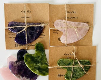 May include: Four Gua Sha tools made of different stones: amethyst, rose quartz, jade, and green jade. Each tool is tied with twine and packaged on a brown card with the name of the stone and the text "Gua Sha".
