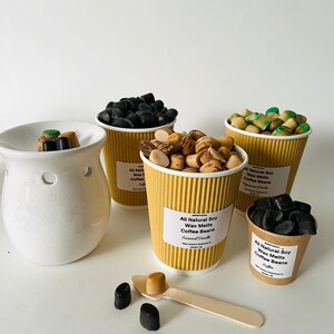 May include: Four brown paper cups filled with soy wax melts in various scents, including coffee beans, caramel vanilla, and peppermint mocha. The wax melts are in the shape of small coffee beans. A white ceramic wax warmer is in the background.