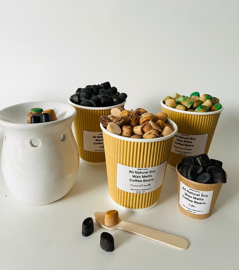 May include: Four brown paper cups filled with soy wax melts in various scents, including coffee beans, caramel vanilla, and peppermint mocha. The wax melts are in the shape of small coffee beans. A white ceramic wax warmer is in the background.