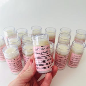 May include: A close-up of a hand holding a clear plastic tube of solid body lotion. The label reads "Solid Body Lotion Warm Vanilla Scent Nourishing/Moisturizing Skincare". There are multiple tubes of lotion in the background.