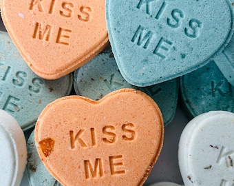 May include: Close-up of heart-shaped candies in shades of orange, blue, and white. The orange candies have the text "KISS ME" imprinted on them. The blue candies also have the text "KISS ME". The candies are a Valentine's Day treat.