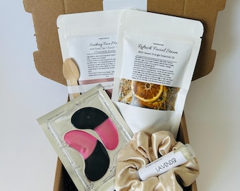 May include: A brown cardboard box filled with self-care products. The box contains two white pouches labeled "Soothing Face Ma" and "Refresh Facial Steam", a wooden spoon, eye patches, a gold scrunchie, and a lip balm labeled "LAVENDER".