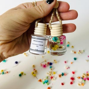 May include: Two small glass bottles with cork tops and dried flowers inside. The bottles are hanging from a string and are labeled "MYBOTANICAL COCONUT CREAM PIE 8 ML".