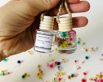 May include: Two small glass bottles with cork tops and dried flowers inside. The bottles are hanging from a string and are labeled "MYBOTANICAL COCONUT CREAM PIE 8 ML".