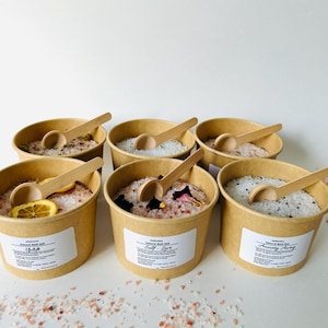 May include: Six small brown paper cups filled with different bath salts. Each cup has a wooden spoon and a label with a name and description. The names are "Uplift", "Self-Love", "Anxiety Away", "Sleep", "Relax", and "Detox".