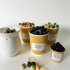 May include: Assortment of coffee bean wax melts in brown paper cups. The wax melts are in various colors, including black, brown, and green. The cups have labels that say "All Natural Soy Wax Melts Coffee Beans".