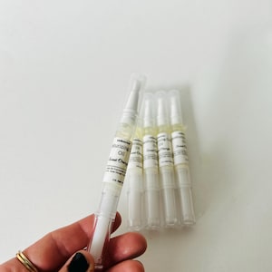 May include: A hand holds a white pen-like tube of moisturizing oil labeled "Sweet Orange." Several similar tubes are arranged on a white surface. The tubes have clear caps and white labels with black text.