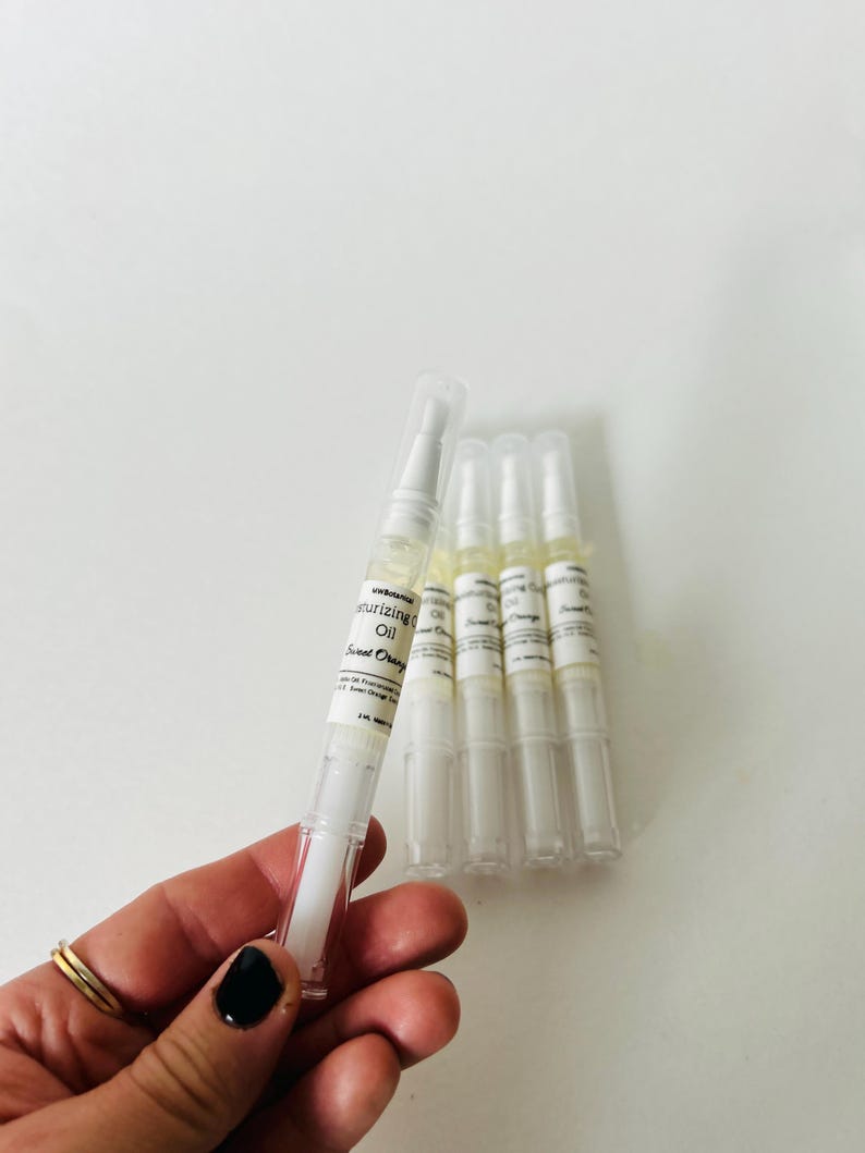 May include: A hand holds a white pen-like tube of moisturizing oil labeled "Sweet Orange." Several similar tubes are arranged on a white surface. The tubes have clear caps and white labels with black text.
