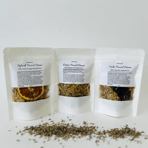 May include: Three white pouches with labels that read "Refresh Facial Steam with Sweet Orange Essential Oil", "Detox Facial Steam with Peppermint Essential Oil", and "Calm Facial Steam with Lavender Essential Oil". Each pouch contains dried herbs and flowers.