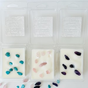 May include: Three clear plastic clamshell containers filled with white soy wax melts. Each container holds different embedded gemstones: bright turquoise stones, pale pink rose quartz, and deep purple amethyst. Loose stones are scattered below.