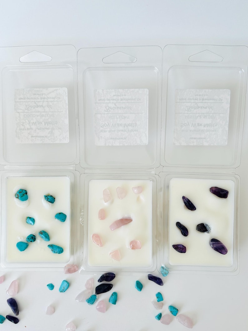 May include: Three clear plastic clamshell containers filled with white soy wax melts. Each container holds different embedded gemstones: bright turquoise stones, pale pink rose quartz, and deep purple amethyst. Loose stones are scattered below.