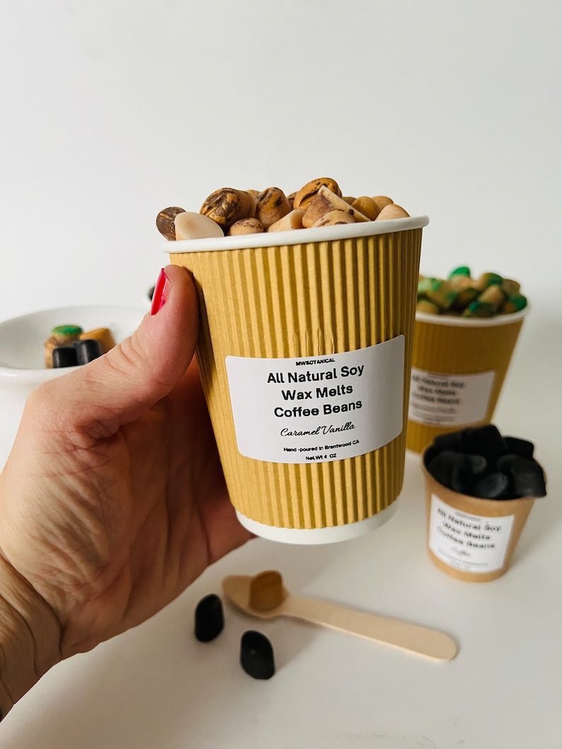 May include: A brown paper cup filled with coffee bean-shaped wax melts. The cup has a label that reads "All Natural Soy Wax Melts Coffee Beans Caramel Vanilla Hand-poured in Brentwood, CA Net Wt. 4 oz."