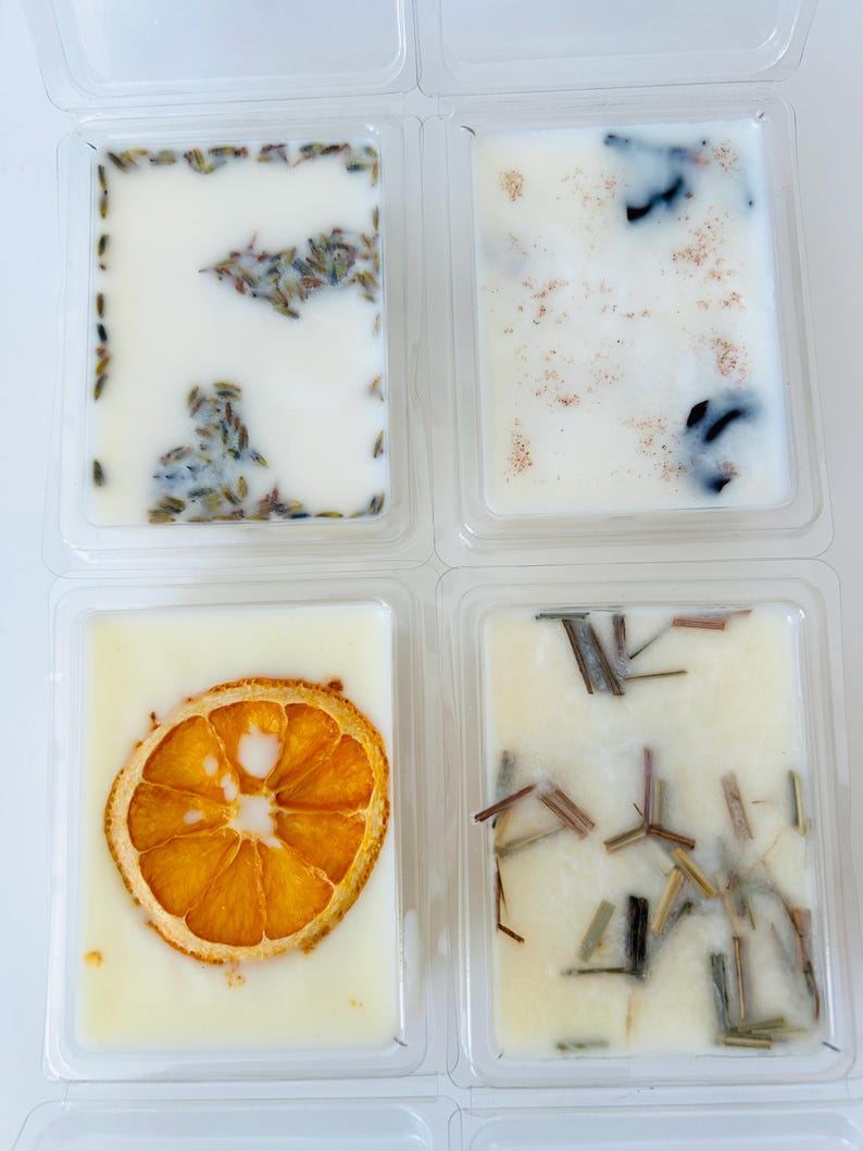 May include: Four rectangular wax melts in clear plastic containers. One contains lavender buds, one has brown spice sprinkles, one features a dried orange slice, and one includes small green botanical stems embedded in the white wax.