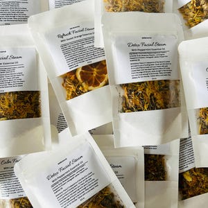 May include: A variety of small white paper pouches filled with dried herbs and flowers. Each pouch has a label with the text "Facial Steam" and the name of an essential oil, such as "Lavender", "Peppermint", or "Sweet Orange".