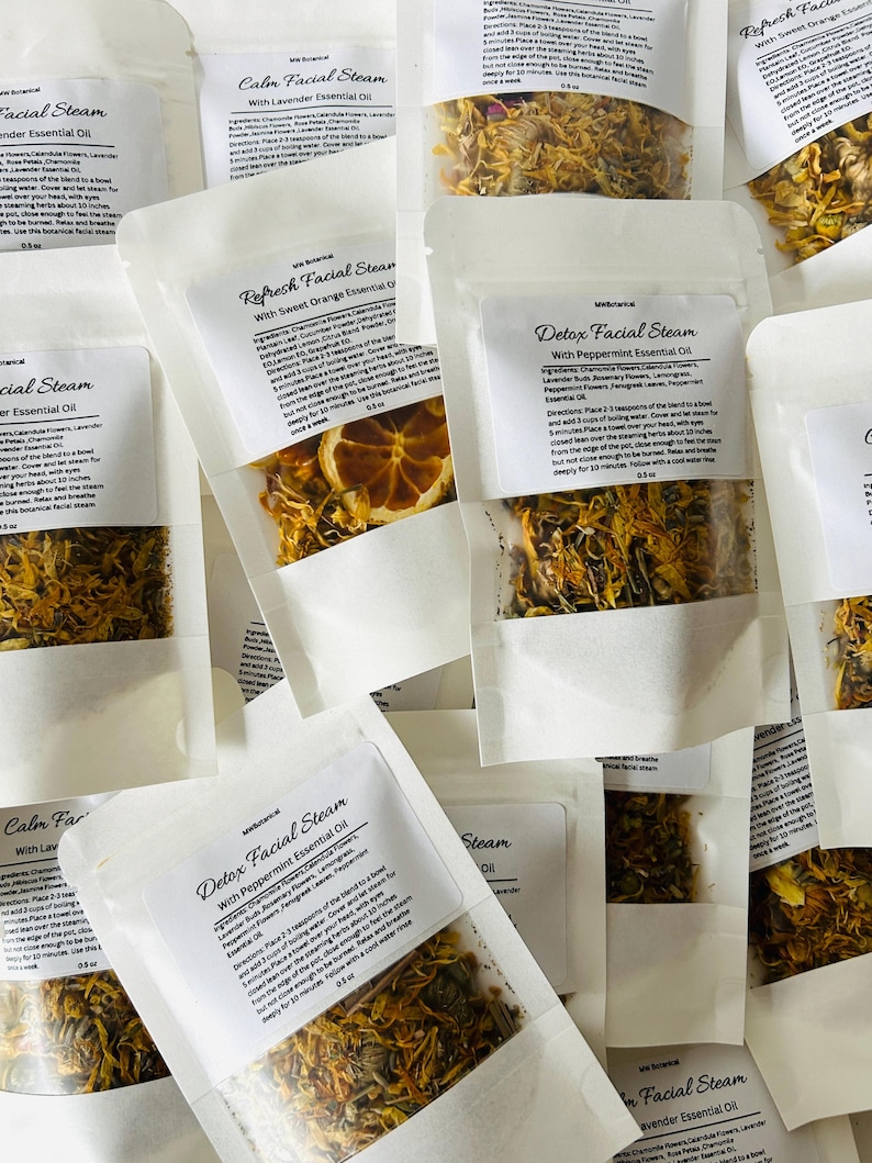 May include: A variety of small white paper pouches filled with dried herbs and flowers. Each pouch has a label with the text "Facial Steam" and the name of an essential oil, such as "Lavender", "Peppermint", or "Sweet Orange".