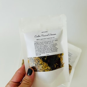 May include: A white pouch with a clear window showing a blend of dried herbs and flowers. The pouch is labeled "Calm Facial Steam with Lavender Essential Oil". The ingredients are listed as chamomile flowers, calendula flowers, lavender buds, hibiscus flowers, rose petals, chamomile powder, and jasmine flowers. The directions for use are also included.