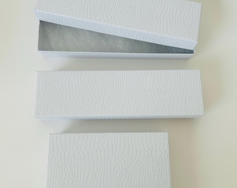 May include: Three white rectangular gift boxes with a textured, embossed surface. One long box is shown open with a white interior lining, while another long box and a smaller rectangular box are shown closed, all featuring a consistent pattern.