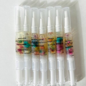 May include: A collection of seven moisturizing cuticle oil pens. Each pen has a clear plastic body with a white cap and contains a colorful oil with floral inclusions. The labels read "Pink Grapeassist", "Dragon Blood Inforest", "Citrus Splash", and "Hazelnut Cal".