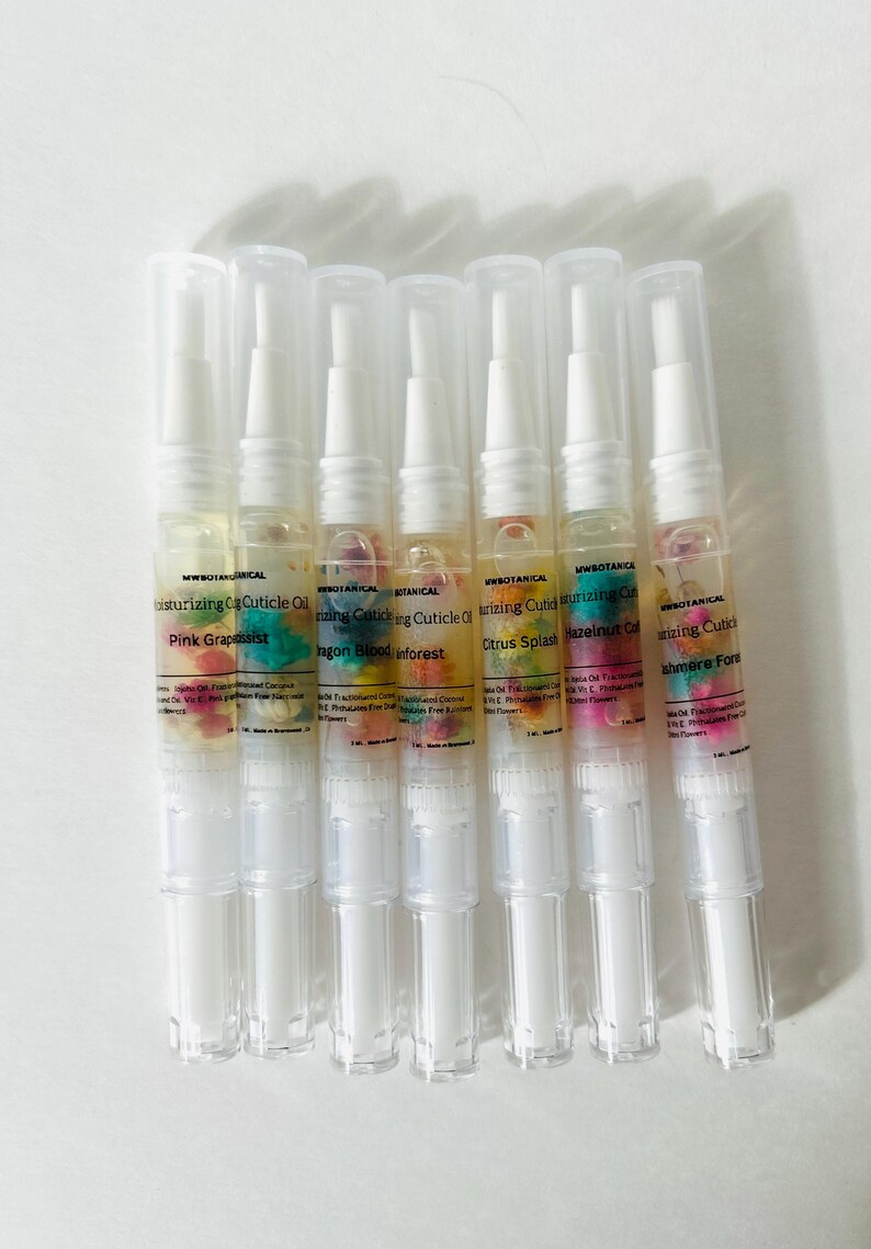 May include: A collection of seven moisturizing cuticle oil pens. Each pen has a clear plastic body with a white cap and contains a colorful oil with floral inclusions. The labels read "Pink Grapeassist", "Dragon Blood Inforest", "Citrus Splash", and "Hazelnut Cal".