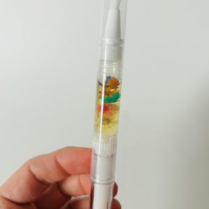 May include: A clear nail cuticle oil pen with a white applicator tip and cap. The pen contains a yellow liquid with colorful dried flowers. The pen is held in a hand against a white background.