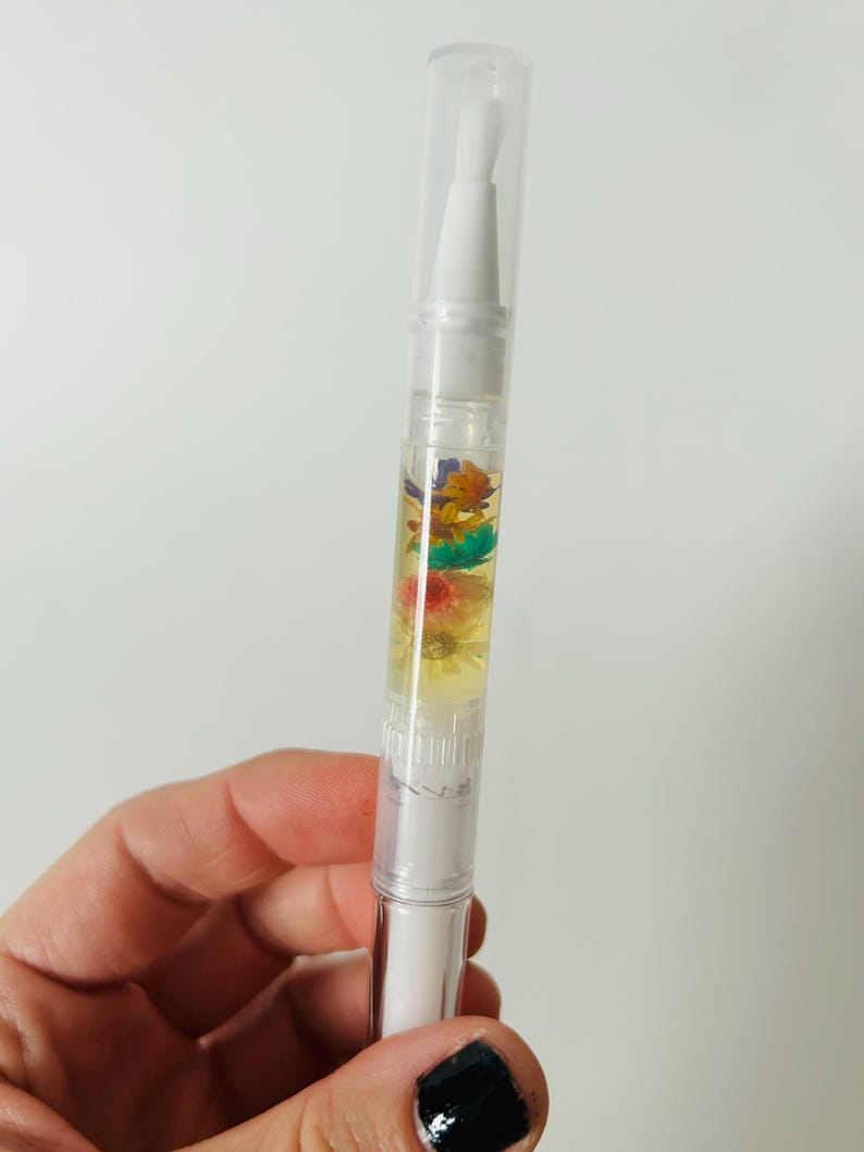 May include: A clear nail cuticle oil pen with a white applicator tip and cap. The pen contains a yellow liquid with colorful dried flowers. The pen is held in a hand against a white background.