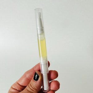 May include: A clear, pen-shaped nail oil applicator with a white brush tip and a yellow liquid inside. The applicator has a clear cap and is held against a white background.
