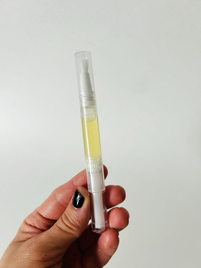 May include: A clear, pen-shaped nail oil applicator with a white brush tip and a yellow liquid inside. The applicator has a clear cap and is held against a white background.