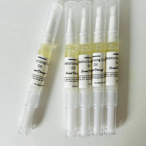 May include: Five clear, pen-shaped containers of moisturizing cuticle oil. The containers have white caps and bases, with a clear section in the middle showing the yellow oil. The label reads "Moisturizing Cuticle Oil Sweet Orange".